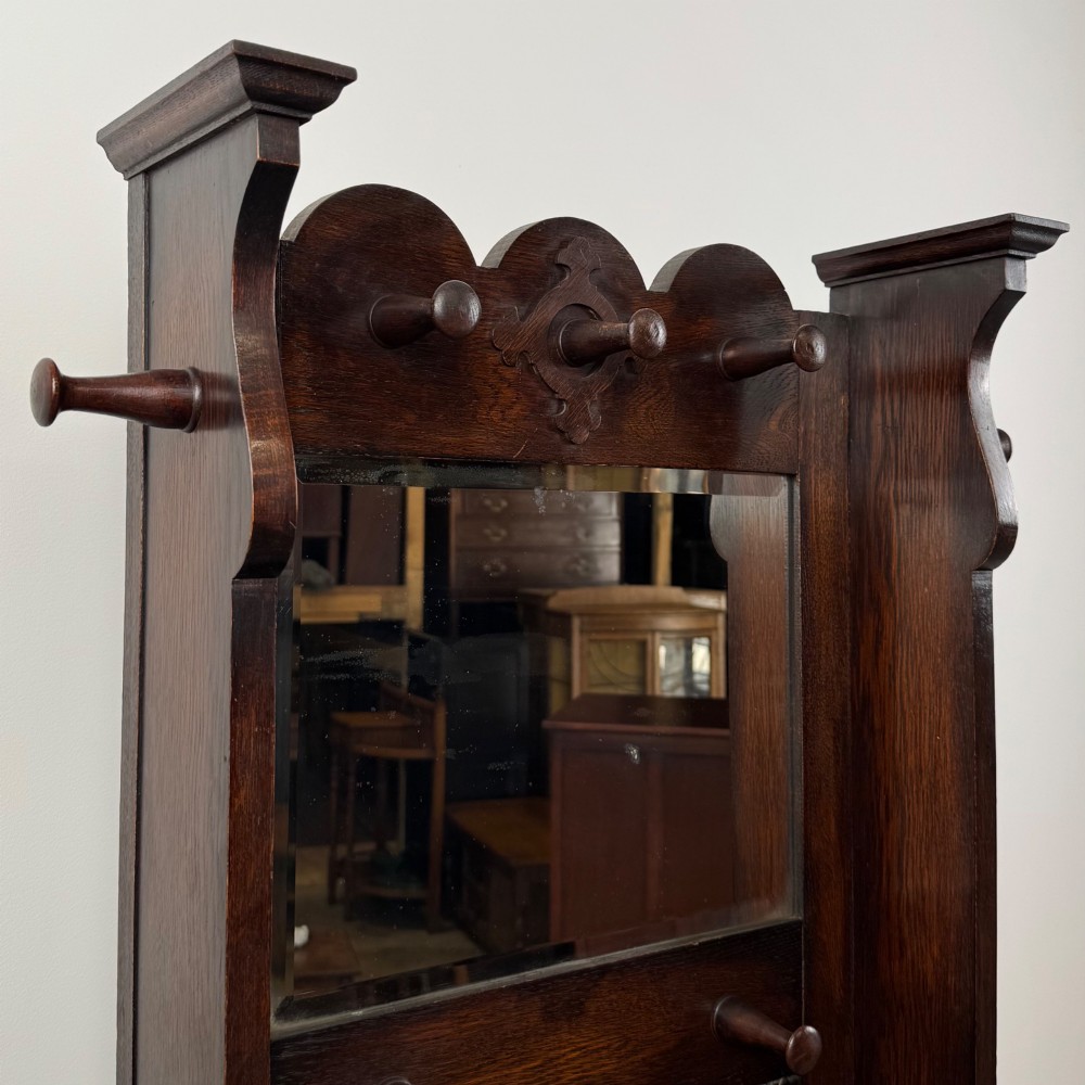 A tall dark oak Edwardian hall stand featuring carved panels, turned coat pegs, a central mirror, a lift-top storage seat, and umbrella holders on both sides. View XII