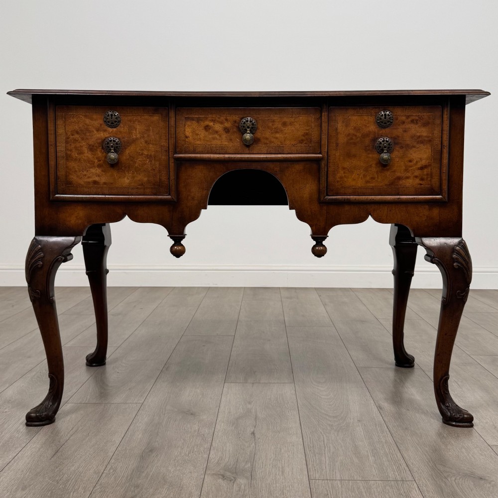 A Queen Anne style burr walnut writing table featuring cabriole legs, three front drawers with brass drop handles, and an arched kneehole.