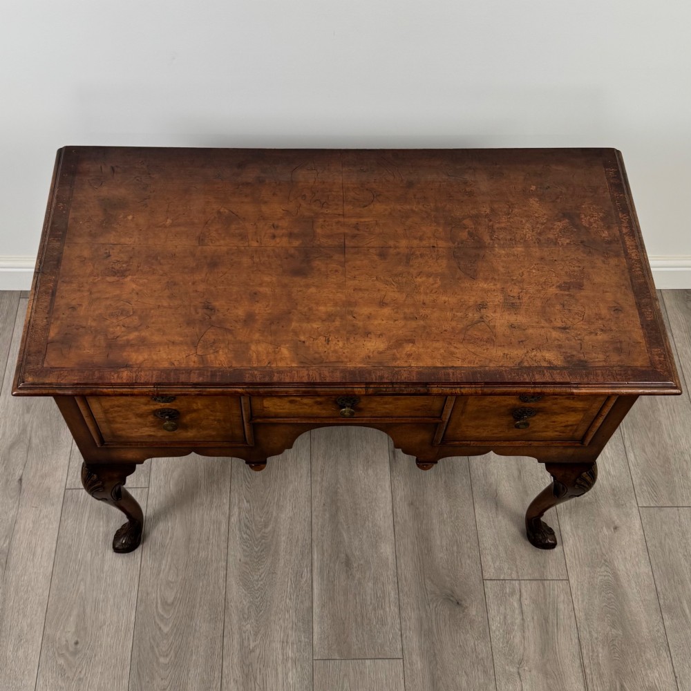 A Queen Anne style burr walnut writing table featuring cabriole legs, three front drawers with brass drop handles, and an arched kneehole. View IX