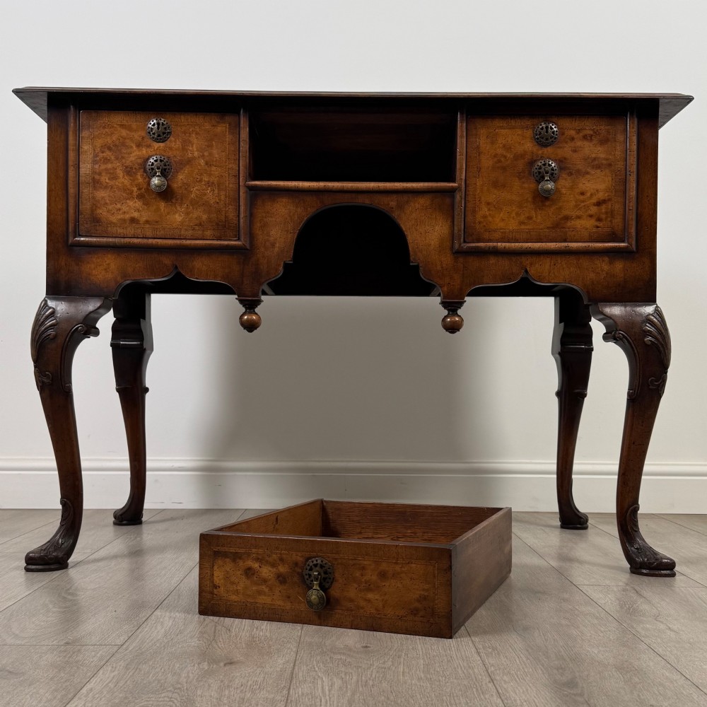 A Queen Anne style burr walnut writing table featuring cabriole legs, three front drawers with brass drop handles, and an arched kneehole. View X