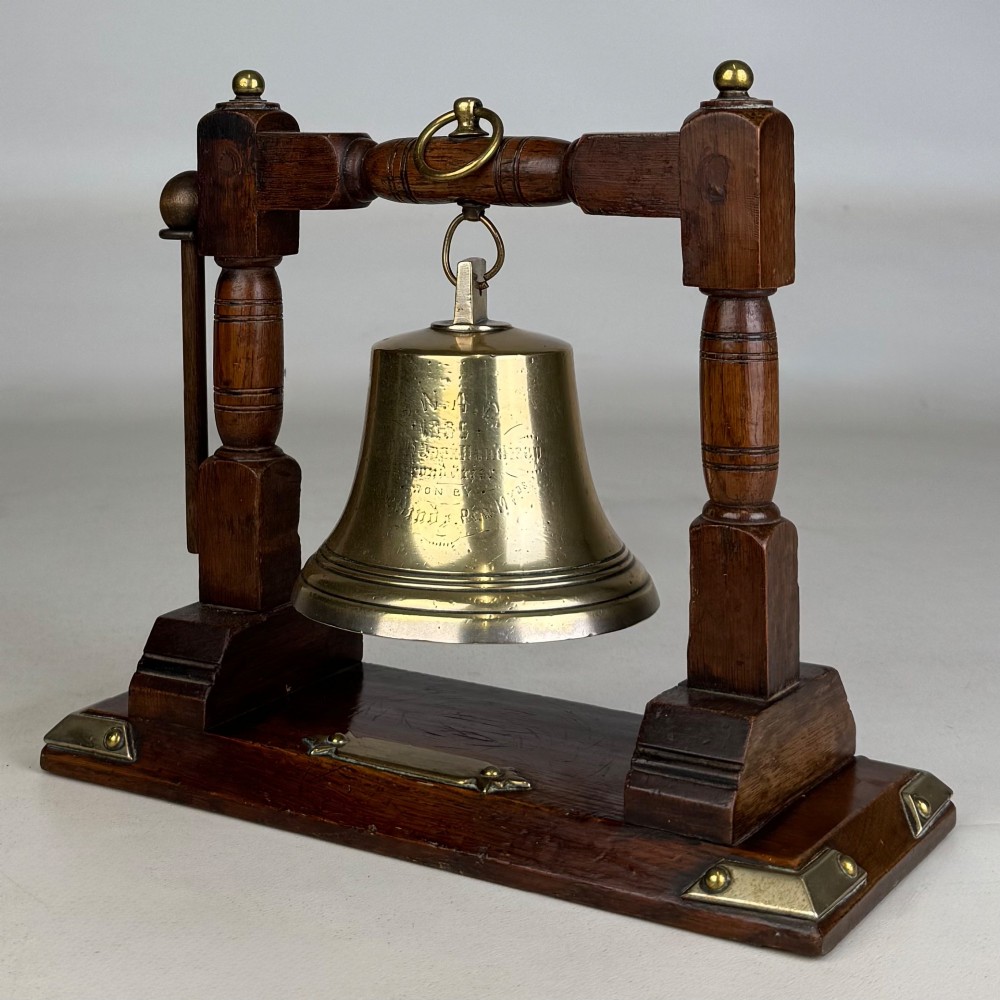 An Edwardian brass bell suspended inside a dark oak wooden frame with turned side pillars, brass corner fittings, and a hanging striker.