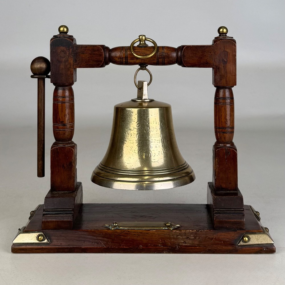 An Edwardian brass bell suspended inside a dark oak wooden frame with turned side pillars, brass corner fittings, and a hanging striker. View II