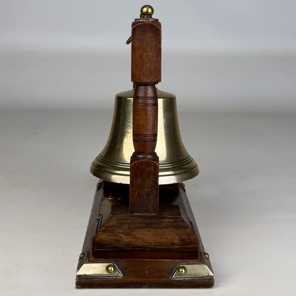 An Edwardian brass bell suspended inside a dark oak wooden frame with turned side pillars, brass corner fittings, and a hanging striker. View III