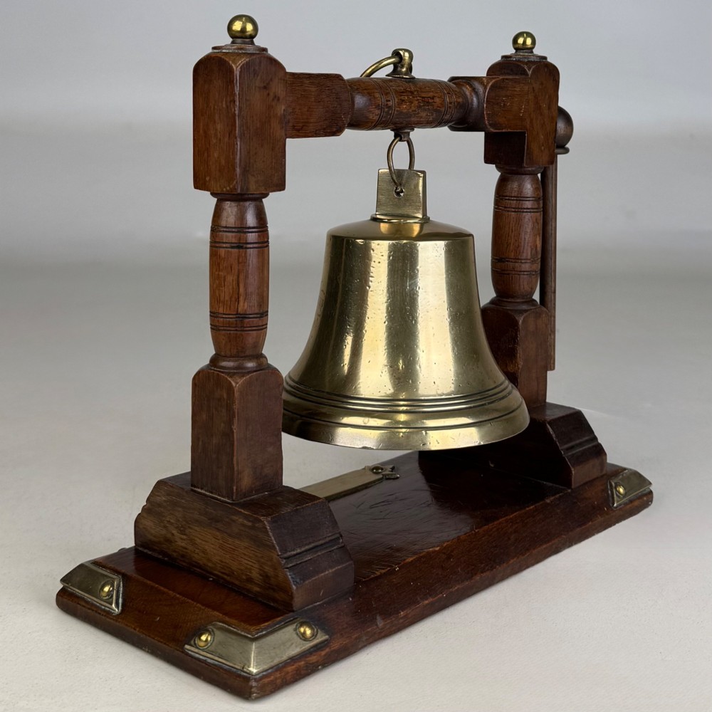 An Edwardian brass bell suspended inside a dark oak wooden frame with turned side pillars, brass corner fittings, and a hanging striker. View IV