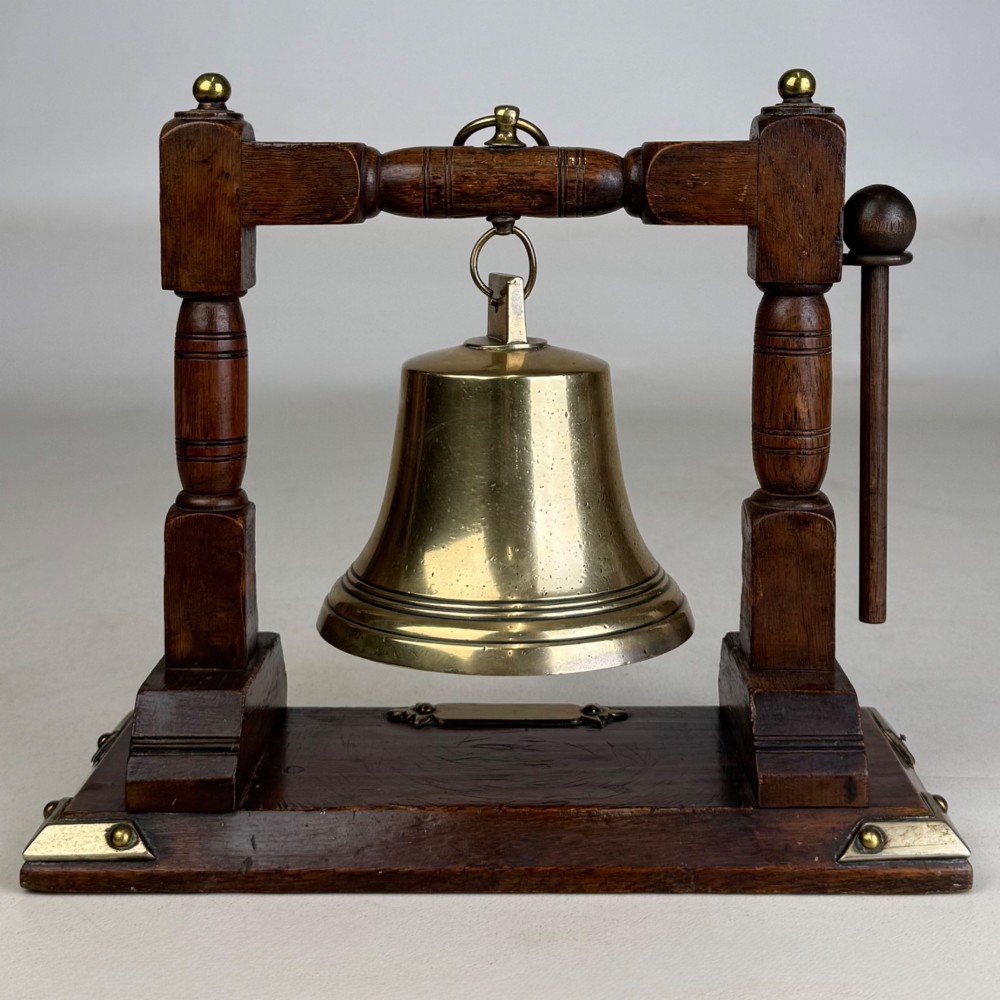 An Edwardian brass bell suspended inside a dark oak wooden frame with turned side pillars, brass corner fittings, and a hanging striker. View V