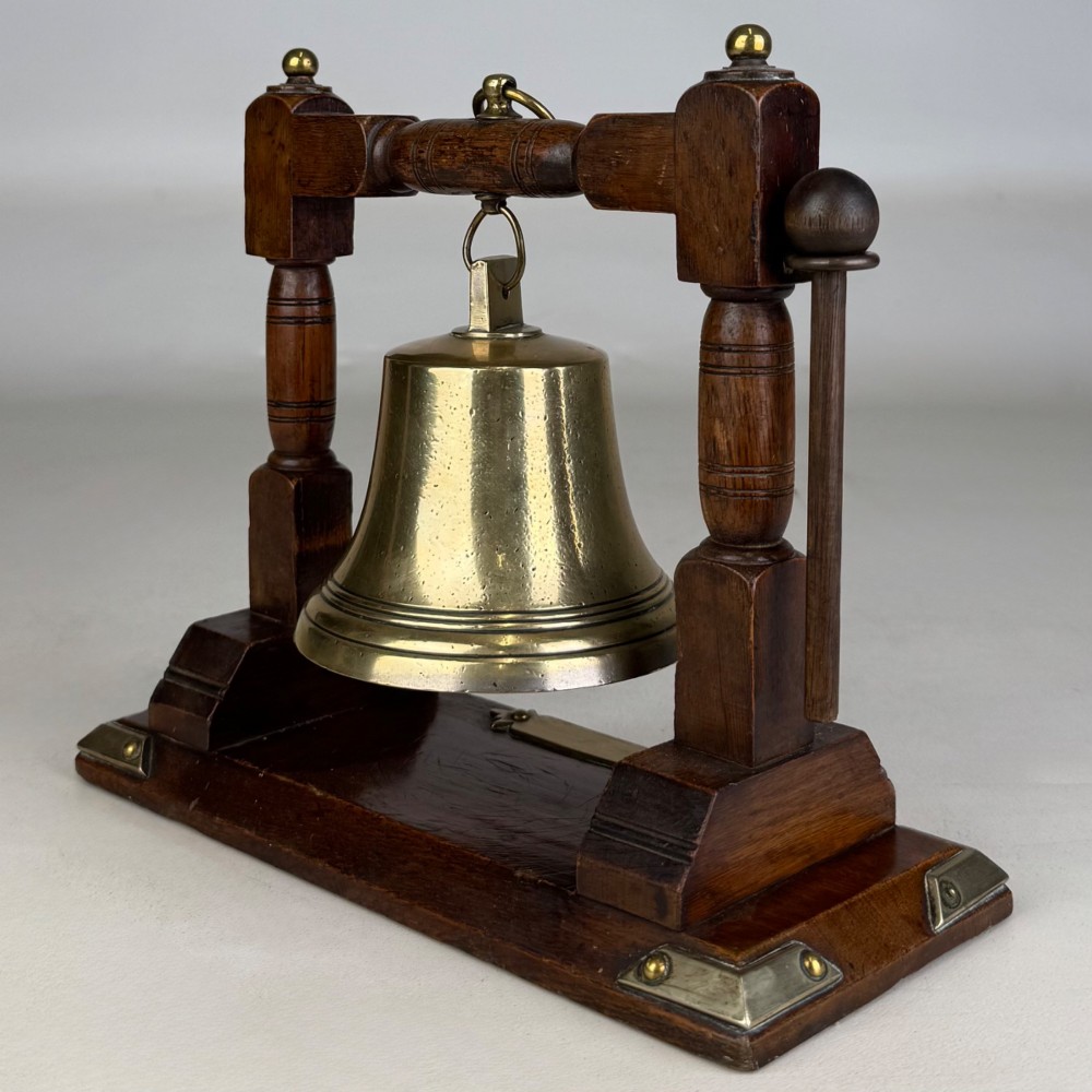 An Edwardian brass bell suspended inside a dark oak wooden frame with turned side pillars, brass corner fittings, and a hanging striker. View VI
