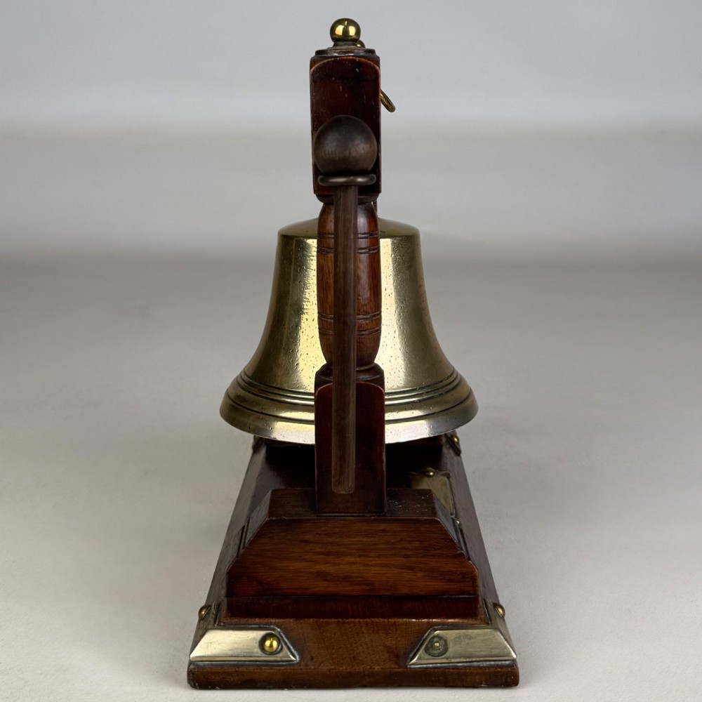An Edwardian brass bell suspended inside a dark oak wooden frame with turned side pillars, brass corner fittings, and a hanging striker. View VII