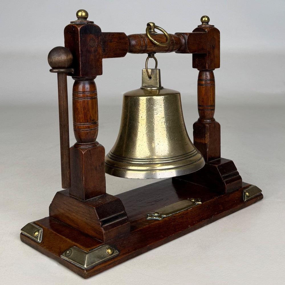 An Edwardian brass bell suspended inside a dark oak wooden frame with turned side pillars, brass corner fittings, and a hanging striker. View VIII