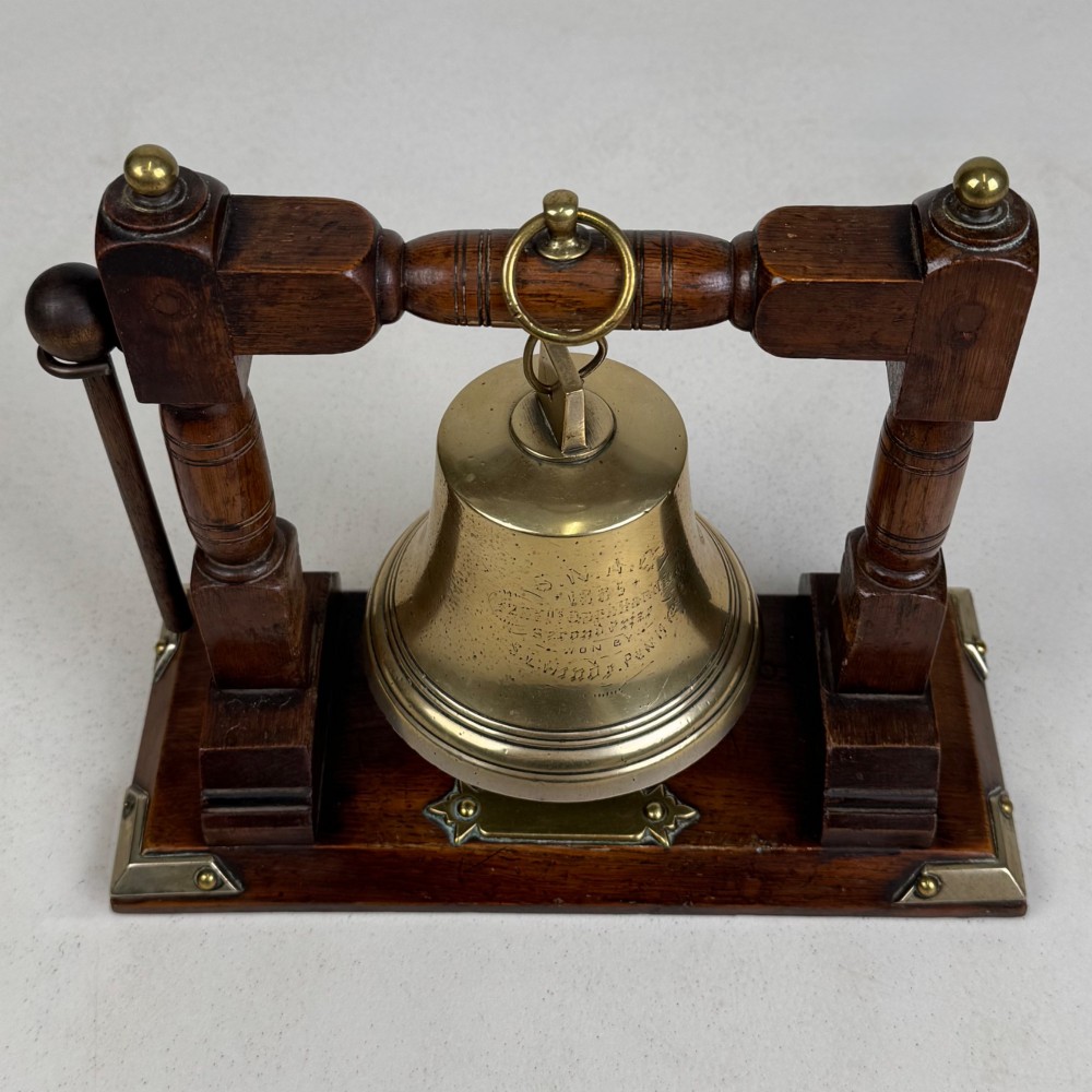 An Edwardian brass bell suspended inside a dark oak wooden frame with turned side pillars, brass corner fittings, and a hanging striker. View IX