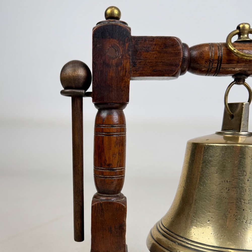 An Edwardian brass bell suspended inside a dark oak wooden frame with turned side pillars, brass corner fittings, and a hanging striker. View X