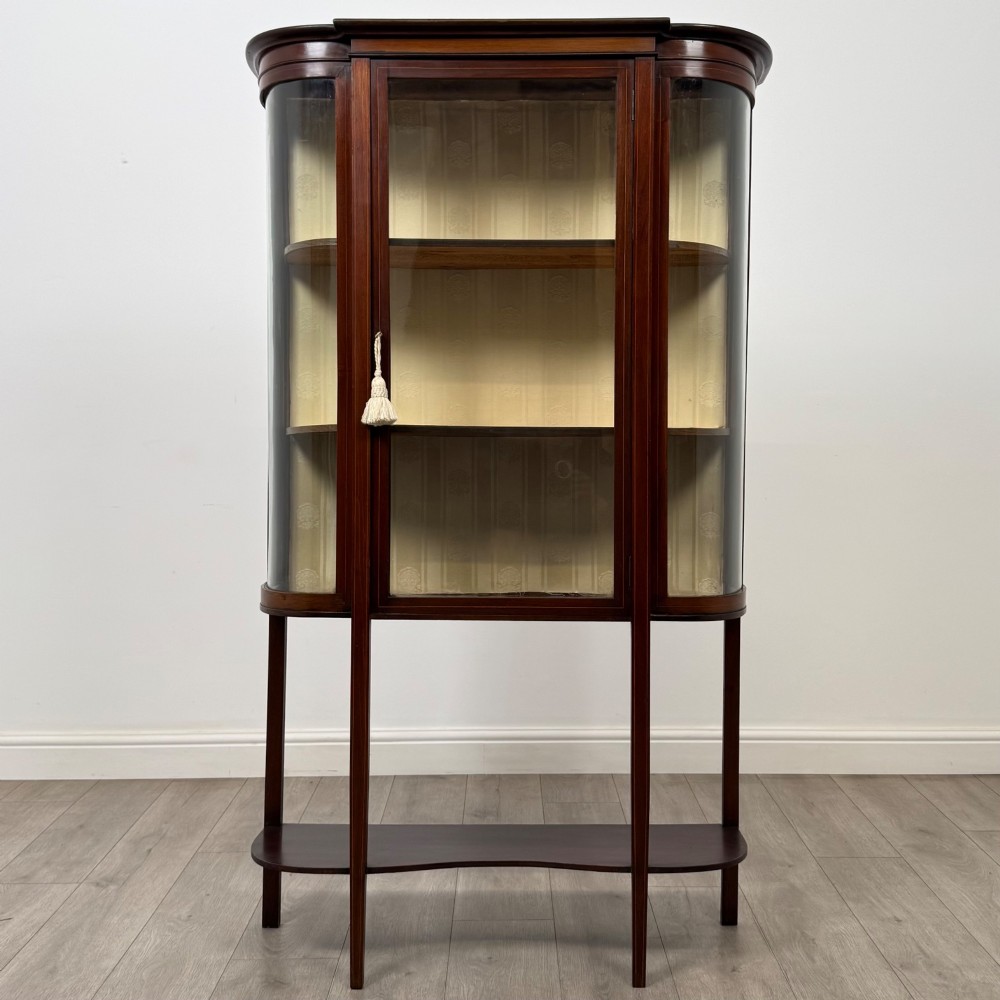 An antique Edwardian mahogany glazed display cabinet featuring curved glass sides, a single central door, fabric-lined interior shelves, and tall slender legs connected by a shaped undertier.