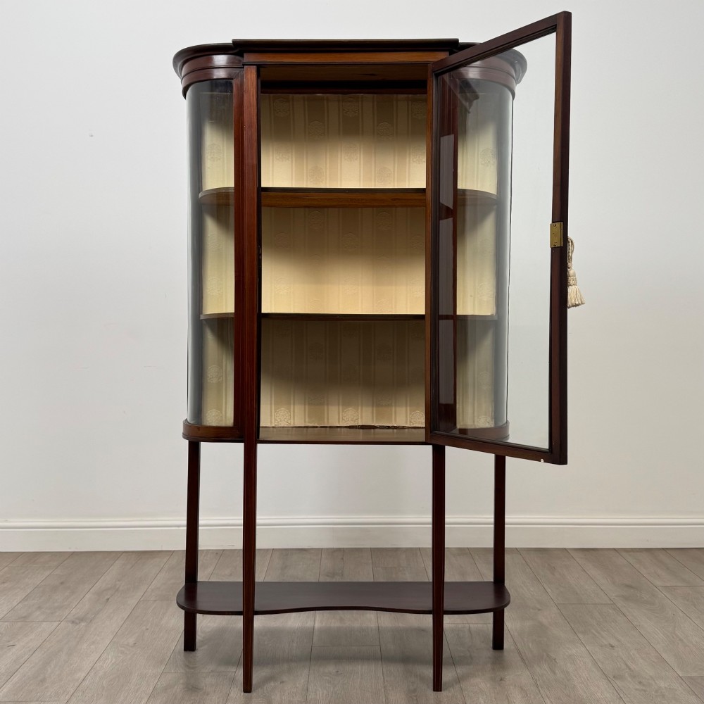 An antique Edwardian mahogany glazed display cabinet featuring curved glass sides, a single central door, fabric-lined interior shelves, and tall slender legs connected by a shaped undertier. View II
