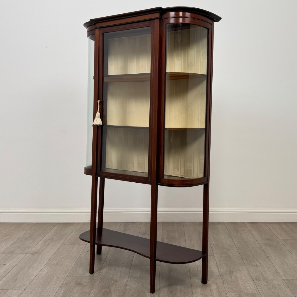An antique Edwardian mahogany glazed display cabinet featuring curved glass sides, a single central door, fabric-lined interior shelves, and tall slender legs connected by a shaped undertier. View III