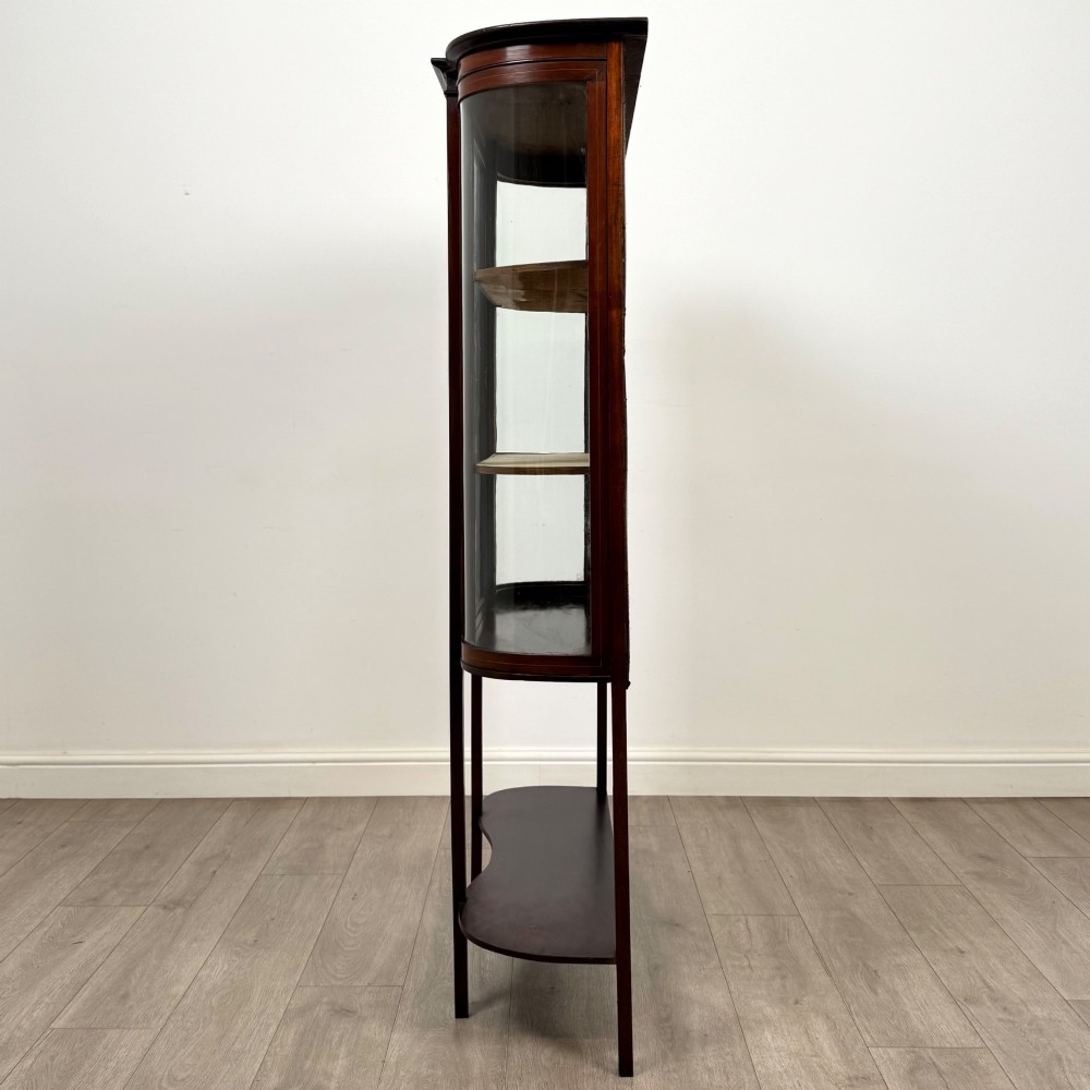 An antique Edwardian mahogany glazed display cabinet featuring curved glass sides, a single central door, fabric-lined interior shelves, and tall slender legs connected by a shaped undertier. View IV