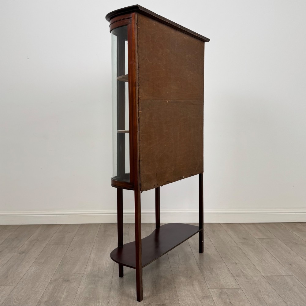 An antique Edwardian mahogany glazed display cabinet featuring curved glass sides, a single central door, fabric-lined interior shelves, and tall slender legs connected by a shaped undertier. View V