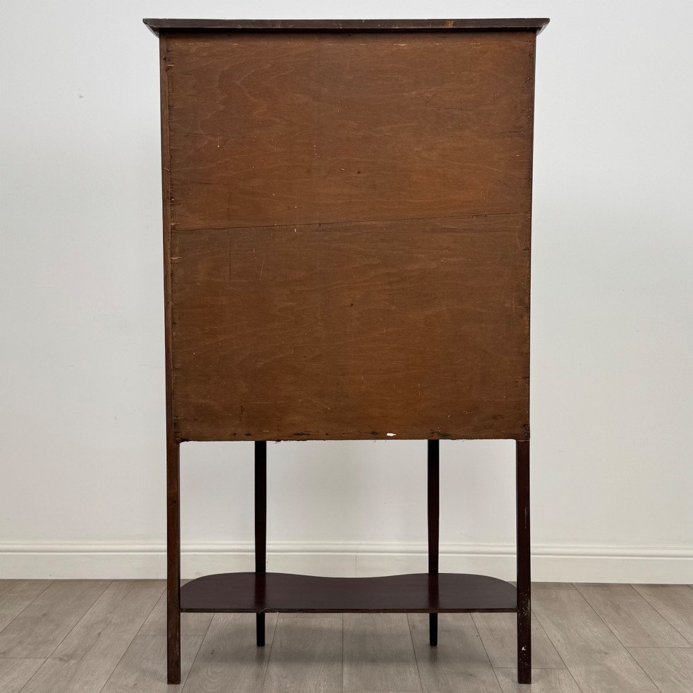 An antique Edwardian mahogany glazed display cabinet featuring curved glass sides, a single central door, fabric-lined interior shelves, and tall slender legs connected by a shaped undertier. View VI