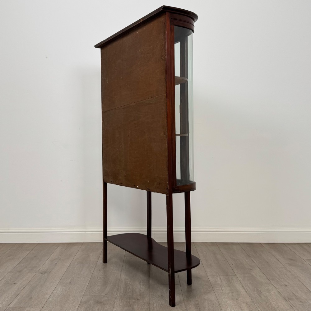 An antique Edwardian mahogany glazed display cabinet featuring curved glass sides, a single central door, fabric-lined interior shelves, and tall slender legs connected by a shaped undertier. View VII