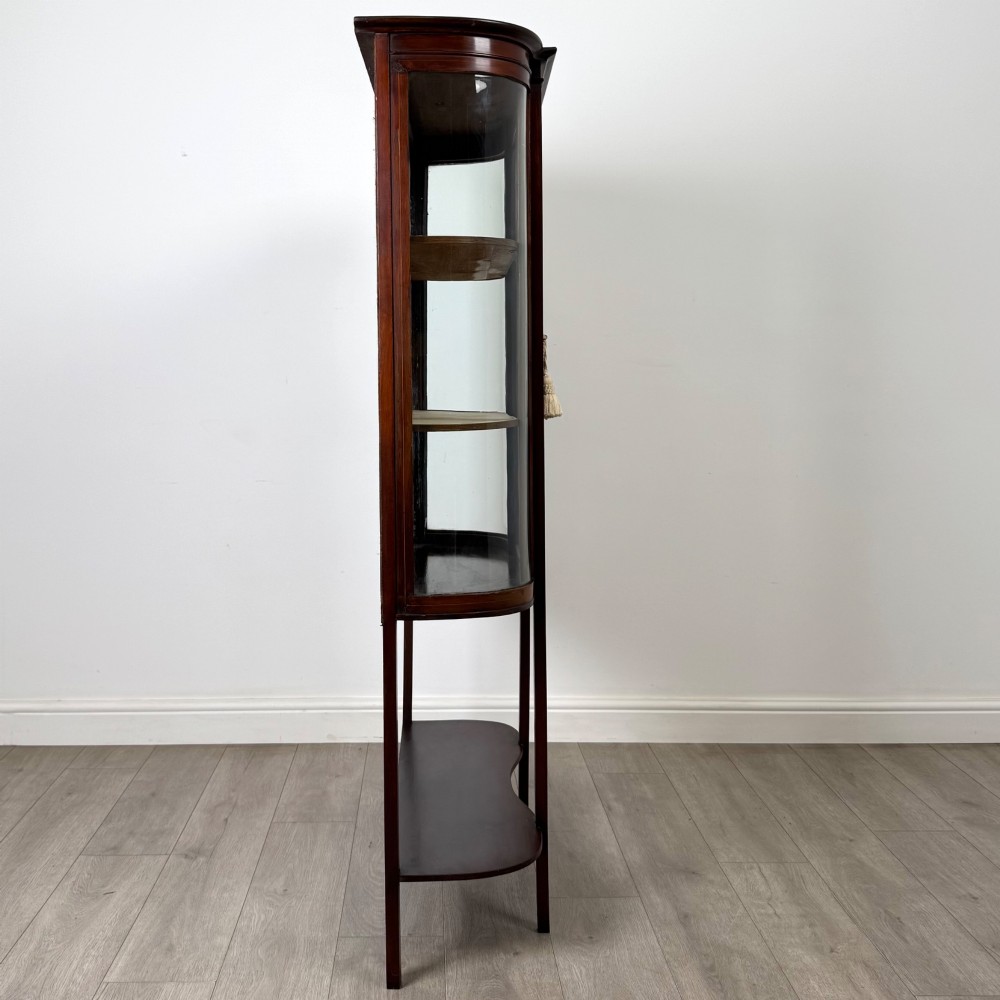 An antique Edwardian mahogany glazed display cabinet featuring curved glass sides, a single central door, fabric-lined interior shelves, and tall slender legs connected by a shaped undertier. View VIII