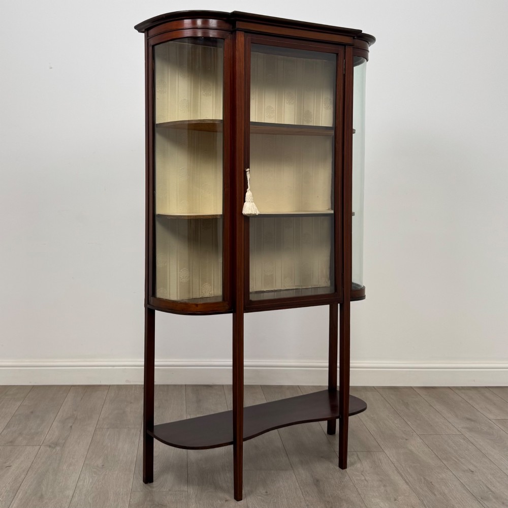 An antique Edwardian mahogany glazed display cabinet featuring curved glass sides, a single central door, fabric-lined interior shelves, and tall slender legs connected by a shaped undertier. View IX