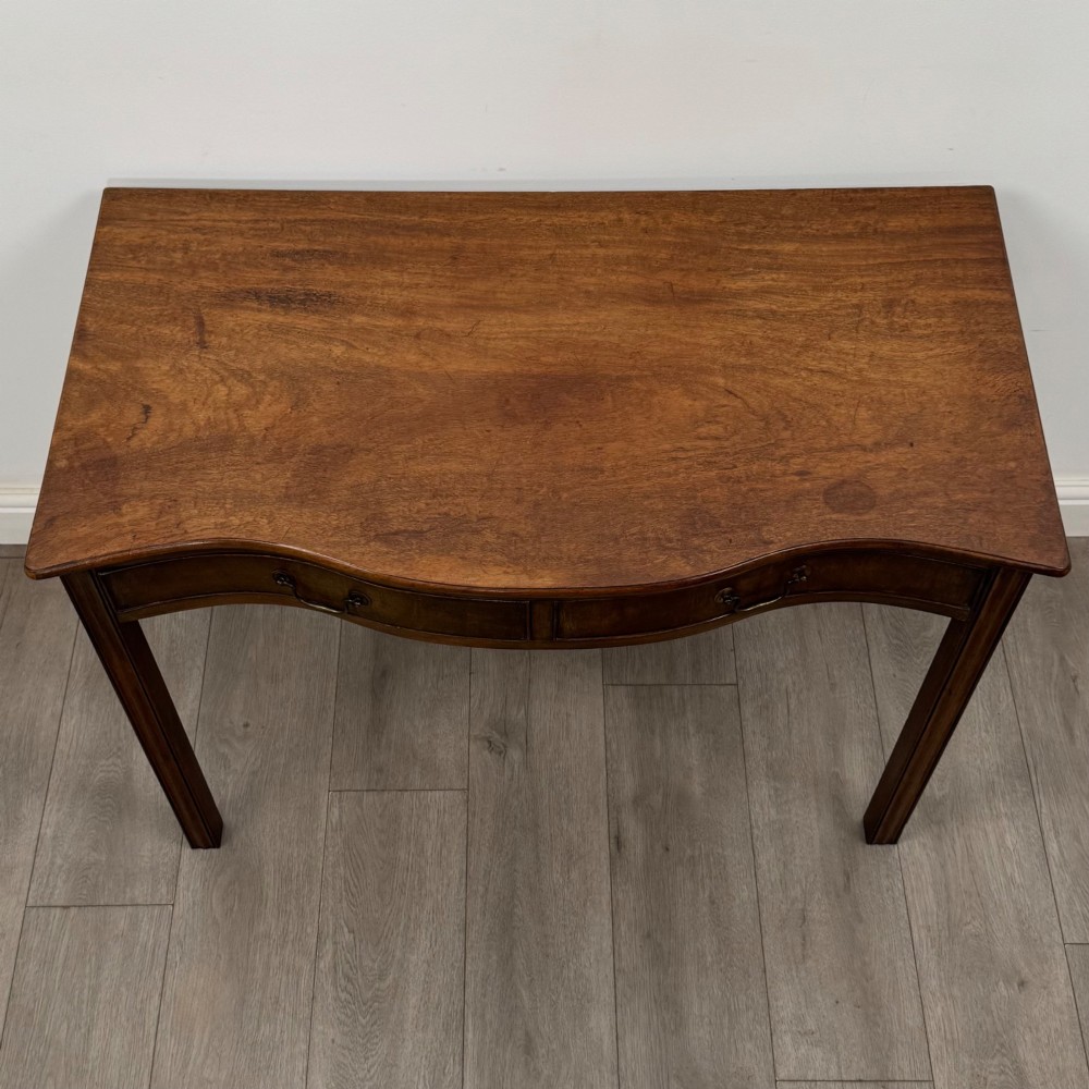 A 19th-century mahogany writing table with a curved serpentine front edge, two drawers with brass handles, and square legs. View IX