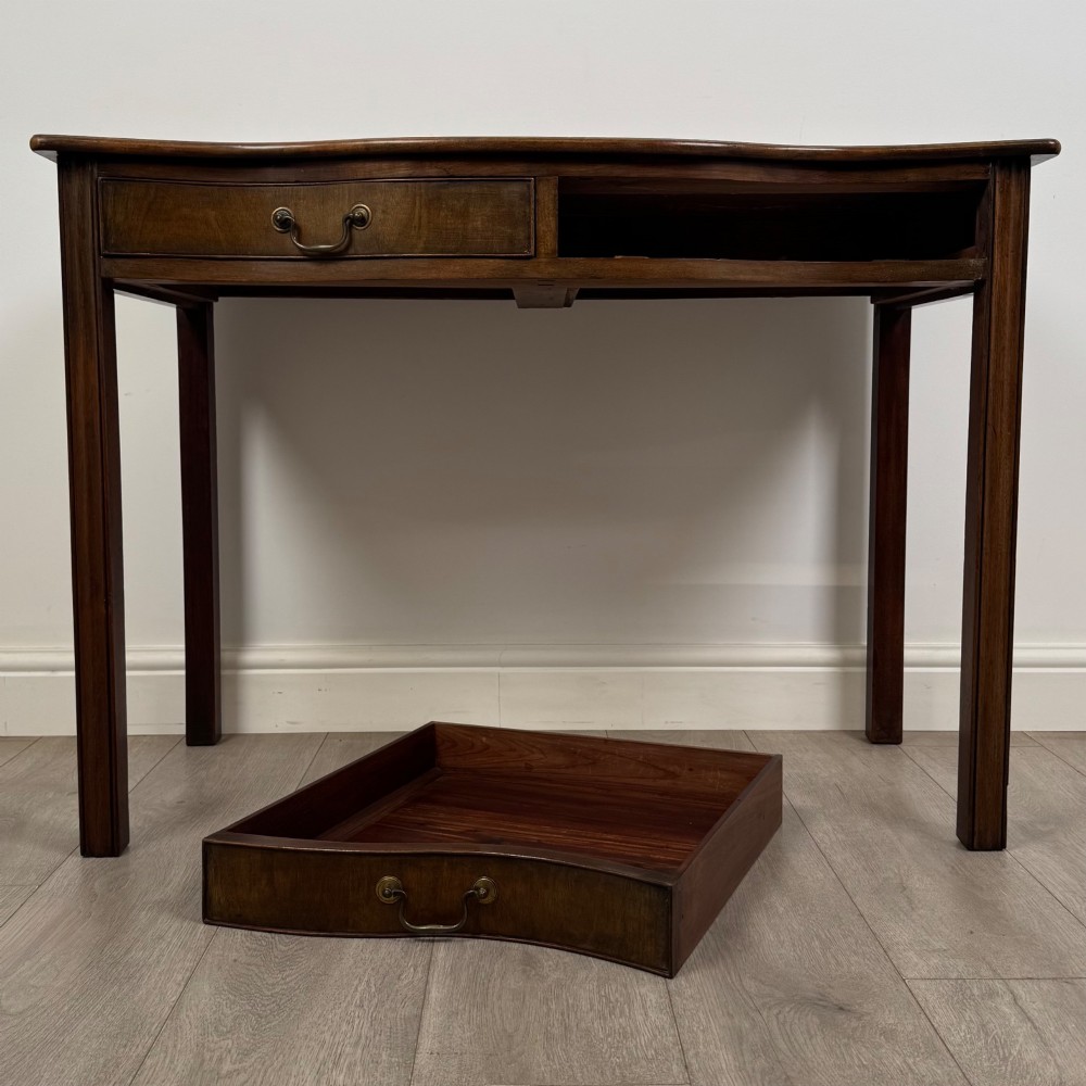 A 19th-century mahogany writing table with a curved serpentine front edge, two drawers with brass handles, and square legs. View X