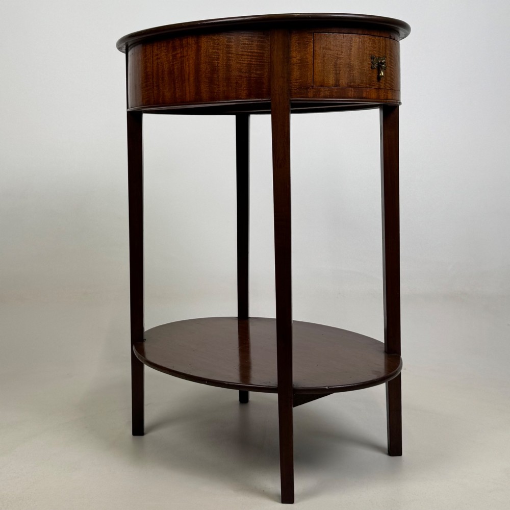 A tall Edwardian mahogany oval side table featuring a single top drawer with a brass handle and an oval lower undertier shelf. View IV