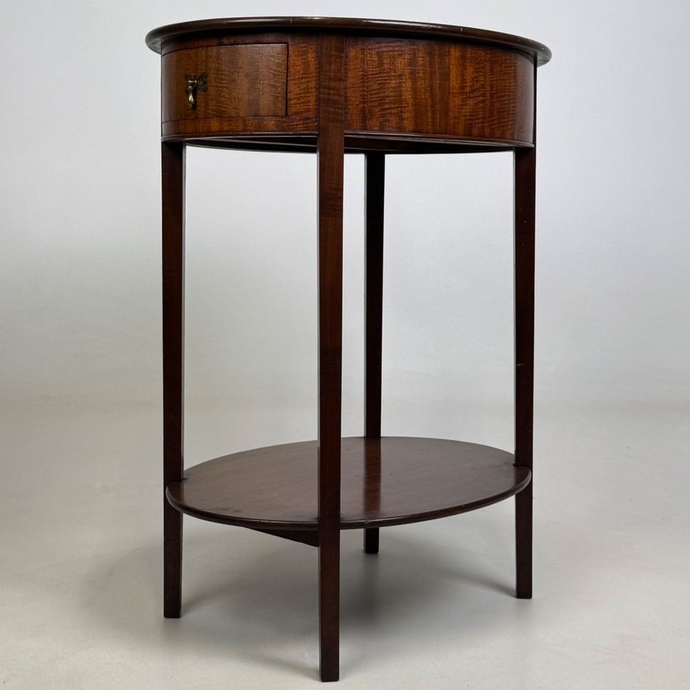 A tall Edwardian mahogany oval side table featuring a single top drawer with a brass handle and an oval lower undertier shelf. View VI
