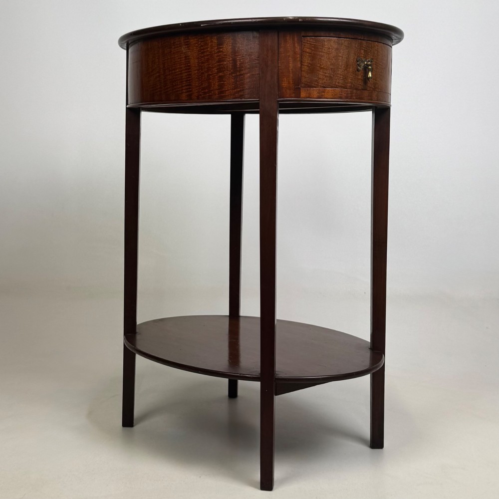 A tall Edwardian mahogany oval side table featuring a single top drawer with a brass handle and an oval lower undertier shelf. View VIII