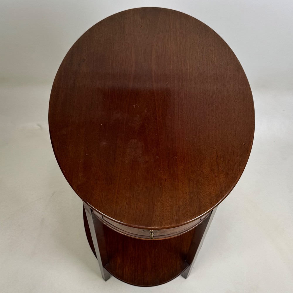 A tall Edwardian mahogany oval side table featuring a single top drawer with a brass handle and an oval lower undertier shelf. View IX