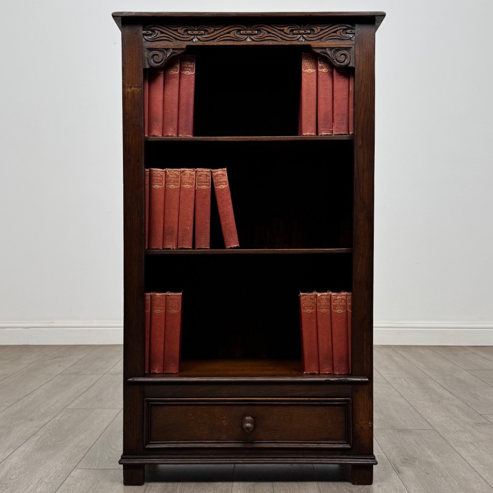 A dark oak open bookcase featuring a carved top frieze, three open display shelves, and a single bottom drawer with a central wooden knob handle.