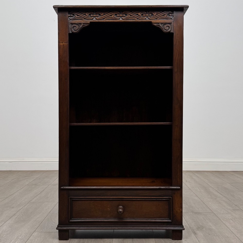 A dark oak open bookcase featuring a carved top frieze, three open display shelves, and a single bottom drawer with a central wooden knob handle. View II