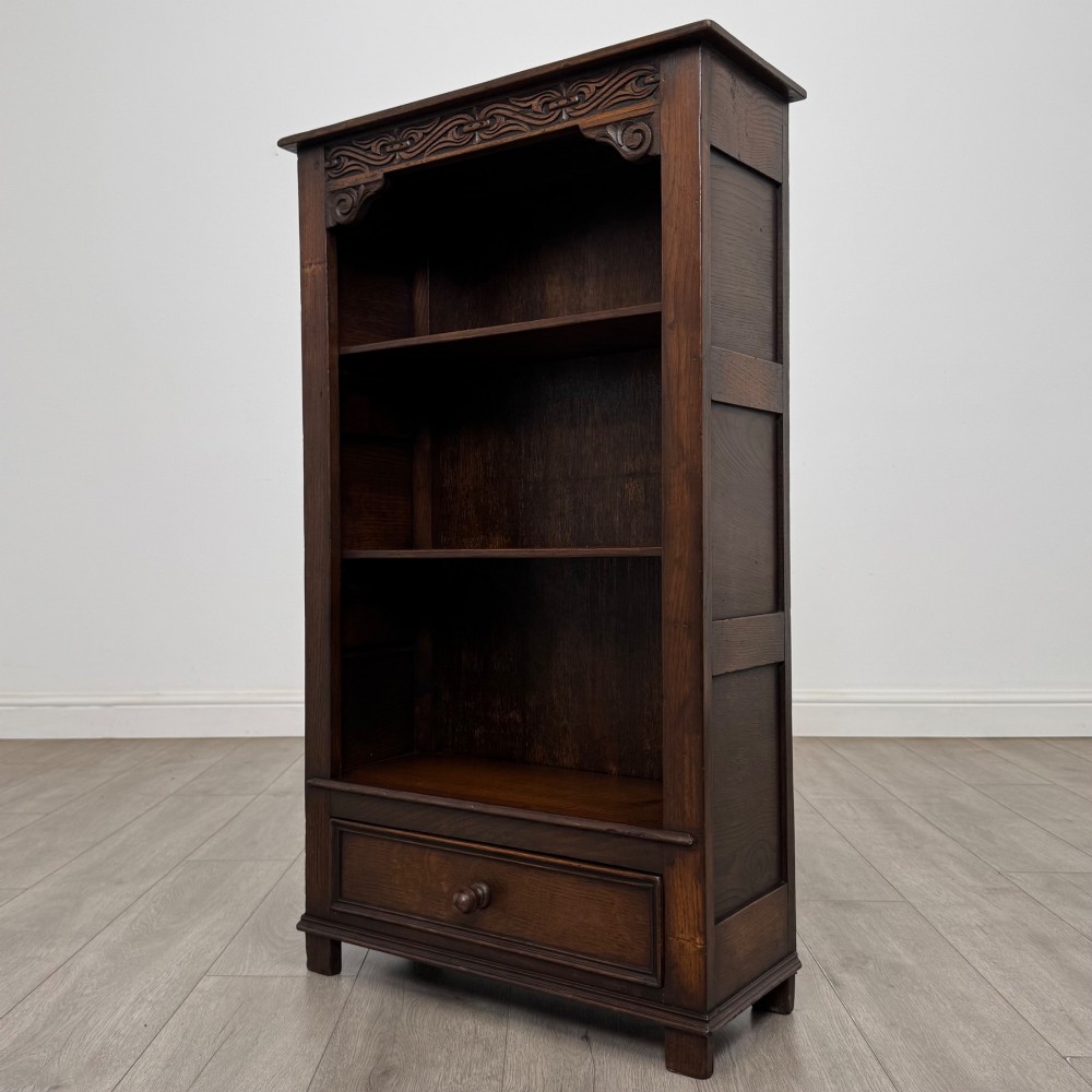 A dark oak open bookcase featuring a carved top frieze, three open display shelves, and a single bottom drawer with a central wooden knob handle. View III
