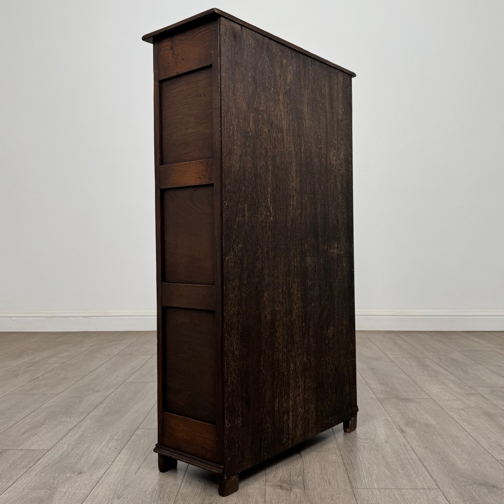 A dark oak open bookcase featuring a carved top frieze, three open display shelves, and a single bottom drawer with a central wooden knob handle. View V