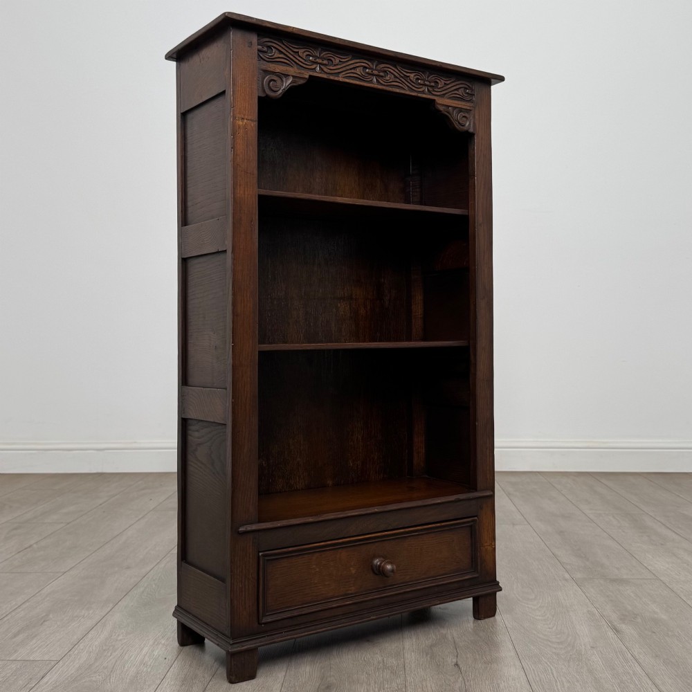 A dark oak open bookcase featuring a carved top frieze, three open display shelves, and a single bottom drawer with a central wooden knob handle. View IX