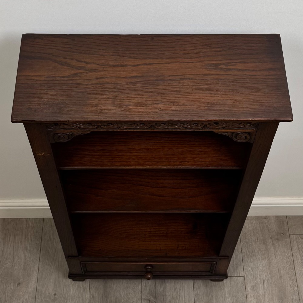 A dark oak open bookcase featuring a carved top frieze, three open display shelves, and a single bottom drawer with a central wooden knob handle. View X