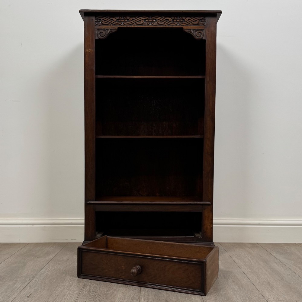 A dark oak open bookcase featuring a carved top frieze, three open display shelves, and a single bottom drawer with a central wooden knob handle. View XI