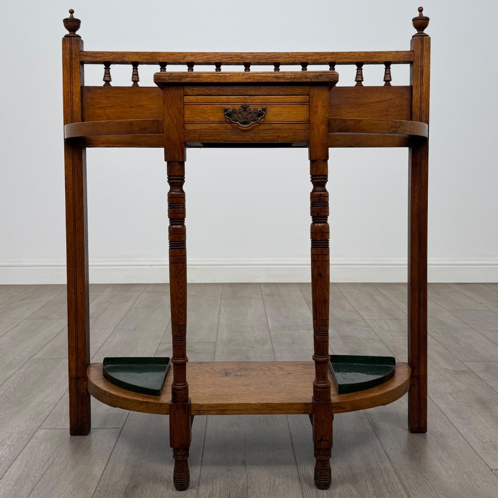 An Edwardian golden oak hall stand with a top spindle gallery, central glove drawer with brass handle, twin umbrella rail apertures, and metal drip trays on the lower shelf.