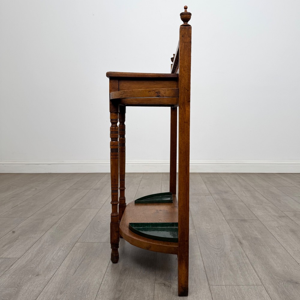 An Edwardian golden oak hall stand with a top spindle gallery, central glove drawer with brass handle, twin umbrella rail apertures, and metal drip trays on the lower shelf. View III