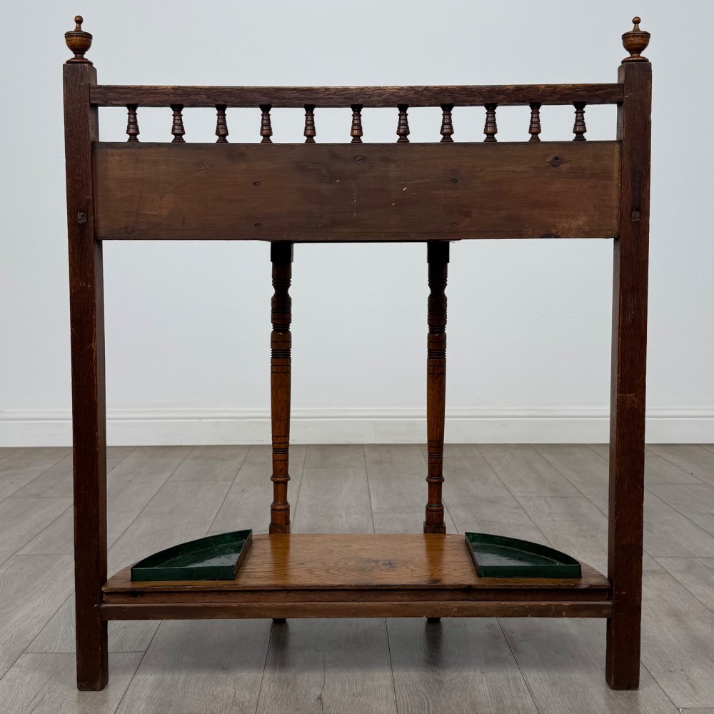 An Edwardian golden oak hall stand with a top spindle gallery, central glove drawer with brass handle, twin umbrella rail apertures, and metal drip trays on the lower shelf. View V