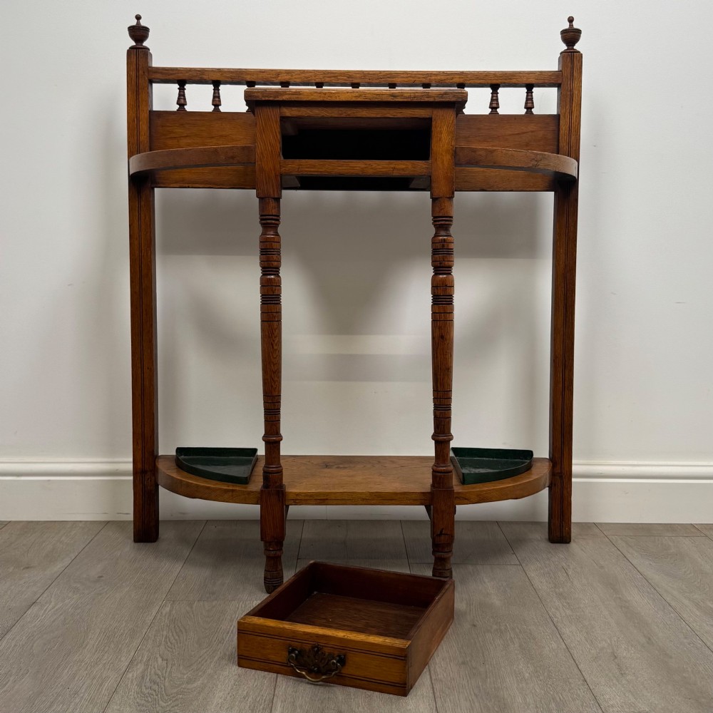 An Edwardian golden oak hall stand with a top spindle gallery, central glove drawer with brass handle, twin umbrella rail apertures, and metal drip trays on the lower shelf. View X