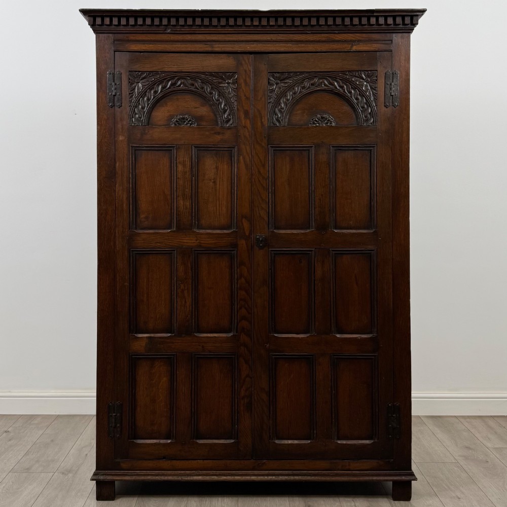 A two-door dark oak wardrobe featuring carved arch top panels, fielded lower door panels, exposed iron strap hinges, and a dentil moulding cornice.