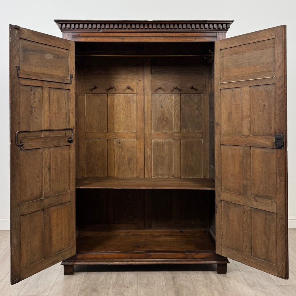 A two-door dark oak wardrobe featuring carved arch top panels, fielded lower door panels, exposed iron strap hinges, and a dentil moulding cornice. View II