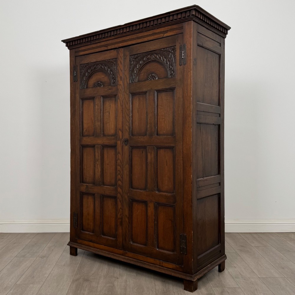 A two-door dark oak wardrobe featuring carved arch top panels, fielded lower door panels, exposed iron strap hinges, and a dentil moulding cornice. View III