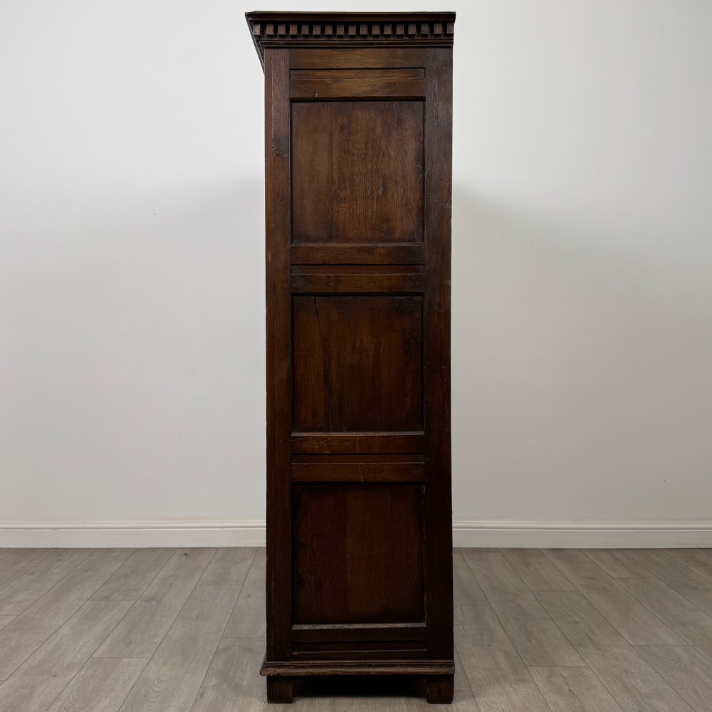 A two-door dark oak wardrobe featuring carved arch top panels, fielded lower door panels, exposed iron strap hinges, and a dentil moulding cornice. View IV
