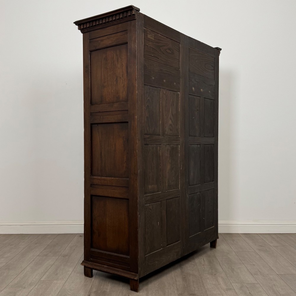 A two-door dark oak wardrobe featuring carved arch top panels, fielded lower door panels, exposed iron strap hinges, and a dentil moulding cornice. View V