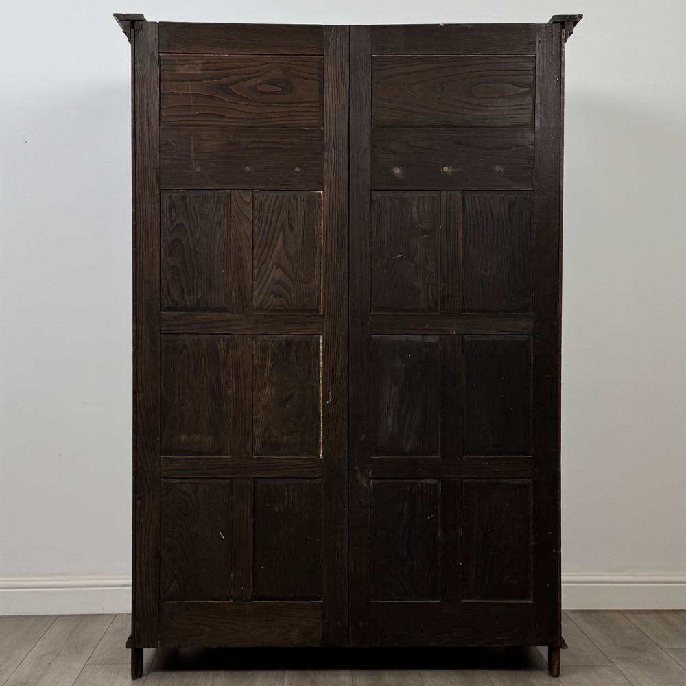 A two-door dark oak wardrobe featuring carved arch top panels, fielded lower door panels, exposed iron strap hinges, and a dentil moulding cornice. View VI