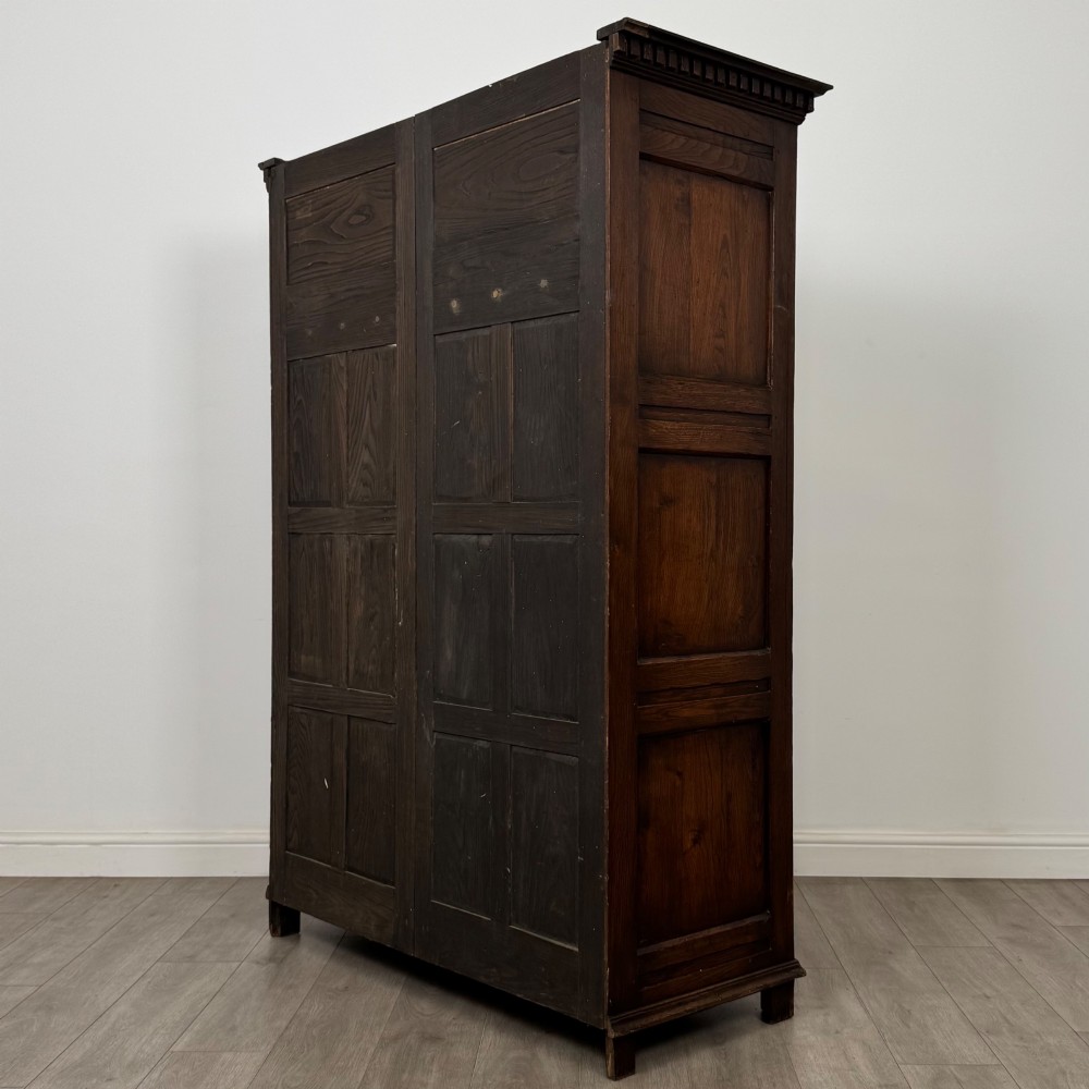 A two-door dark oak wardrobe featuring carved arch top panels, fielded lower door panels, exposed iron strap hinges, and a dentil moulding cornice. View VII