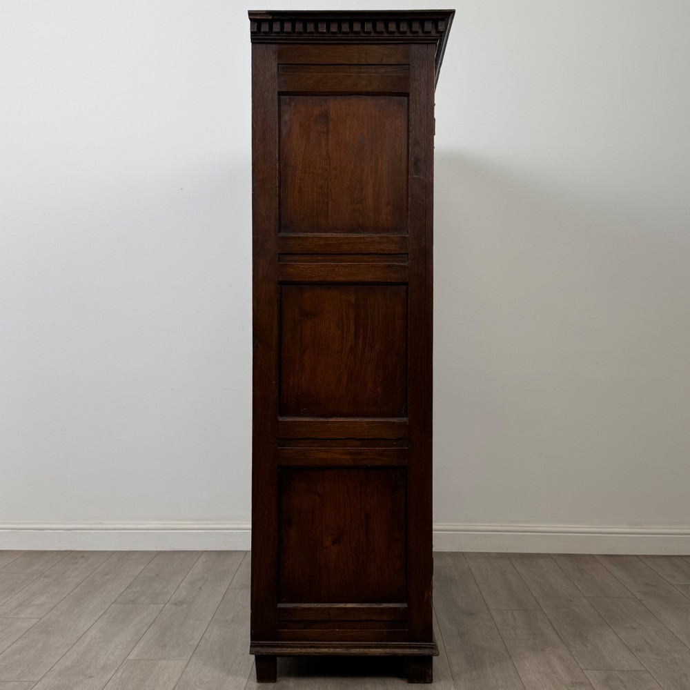 A two-door dark oak wardrobe featuring carved arch top panels, fielded lower door panels, exposed iron strap hinges, and a dentil moulding cornice. View VIII