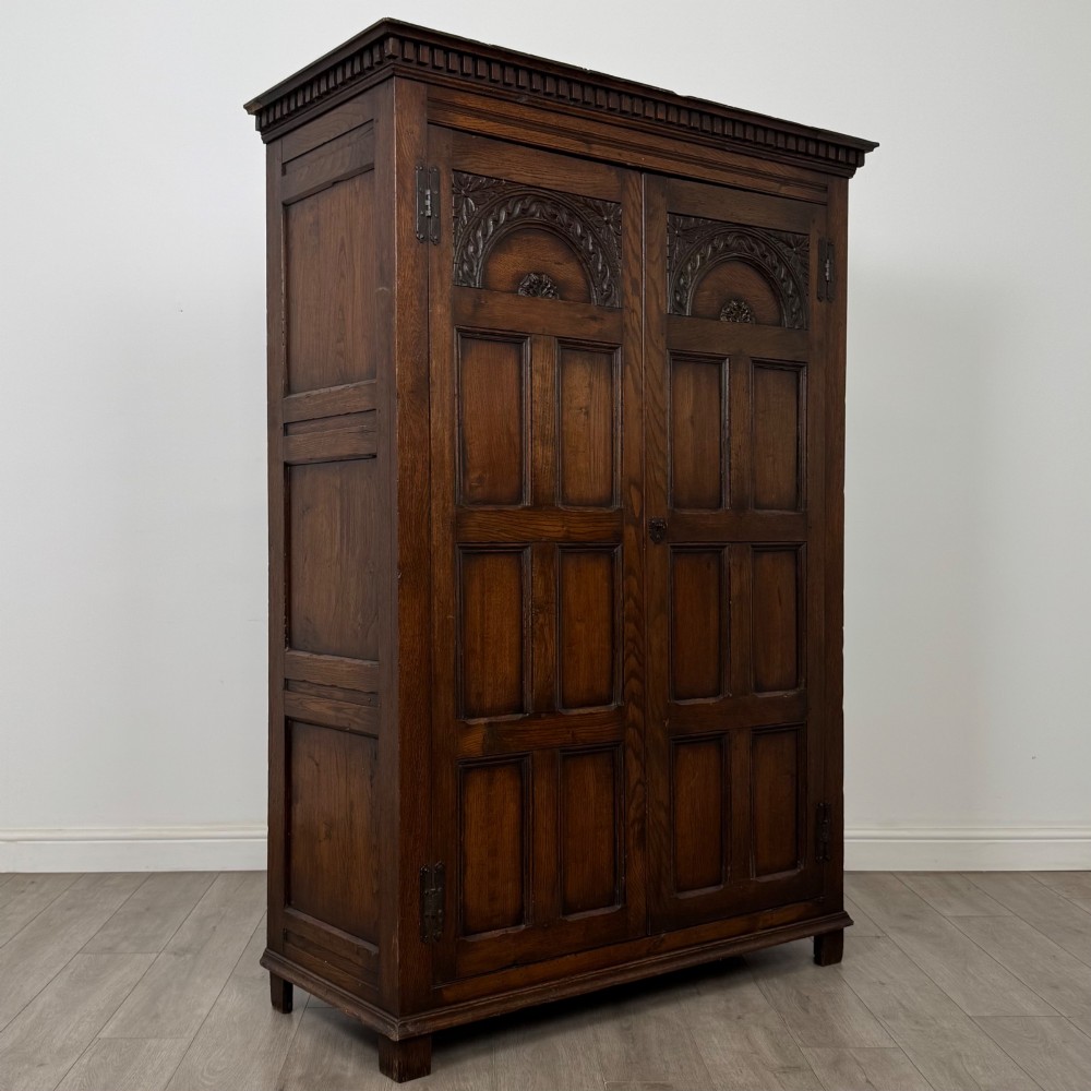A two-door dark oak wardrobe featuring carved arch top panels, fielded lower door panels, exposed iron strap hinges, and a dentil moulding cornice. View IX