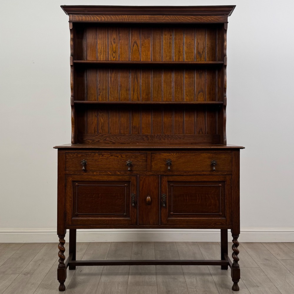 An early 20th-century dark oak Welsh dresser featuring a three-tier open plate rack mounted on a base cabinet with two drawers, geometric panel doors, and barley twist legs.